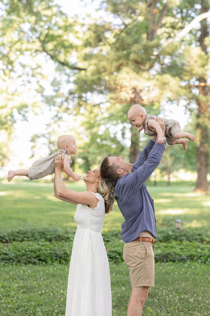 outdoor maternity photography Louisville KY father lifting baby in the air while pregnant mother stands beside him in white dress on green lawn