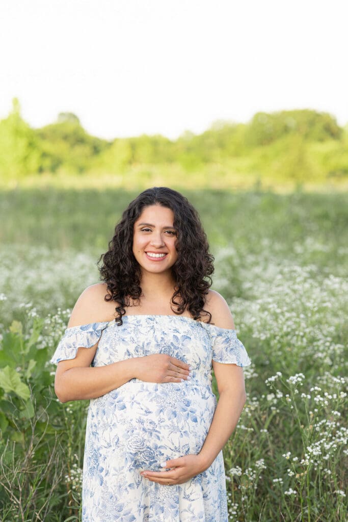maternity photographer Beckley Creek Park Louisville KY pregnant woman in blue floral off shoulder dress holding baby bump in green field at golden hour