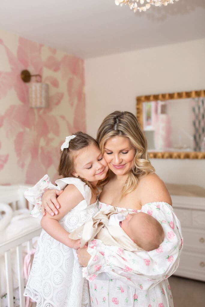 
lifestyle newborn photography Louisville KY mother holding newborn baby girl while older daughter hugs her in pink floral nursery