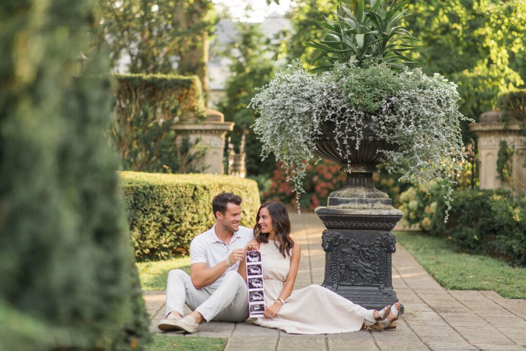 Louisville maternity photographer captures couple sharing ultrasound photos beside ornate garden urn at Whitehall House and Gardens