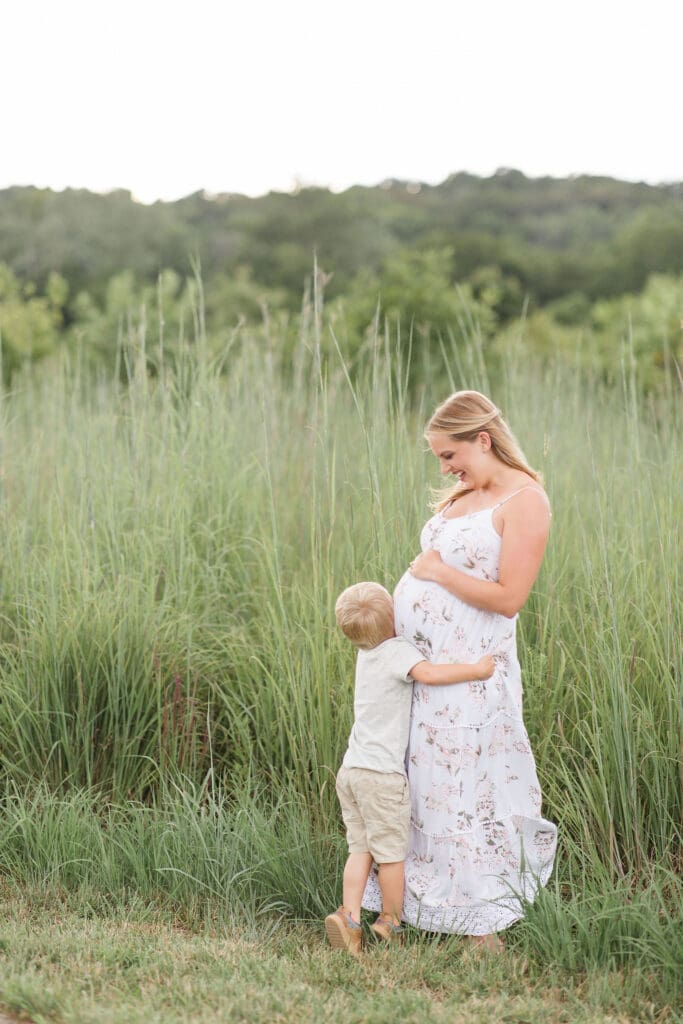 outdoor maternity photography Louisville KY pregnant mother in floral dress smiling at toddler son hugging her baby bump in green field