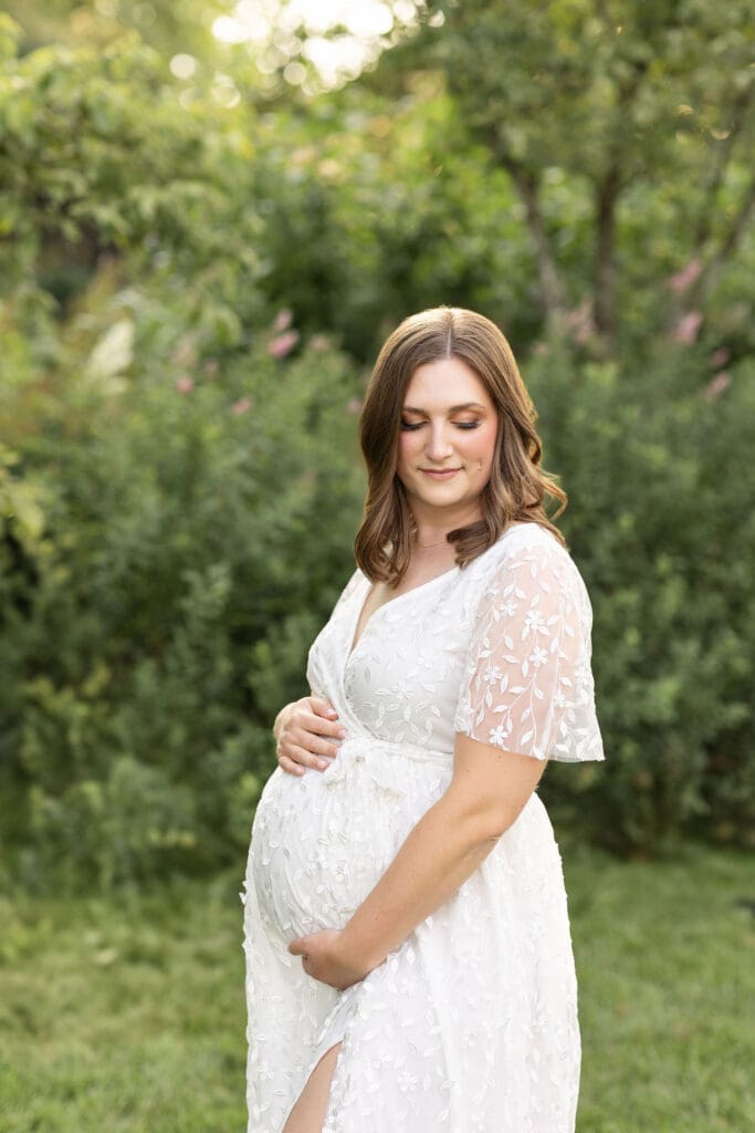 maternity photography Louisville KY pregnant woman in white lace dress holding baby bump outdoors with soft green garden background