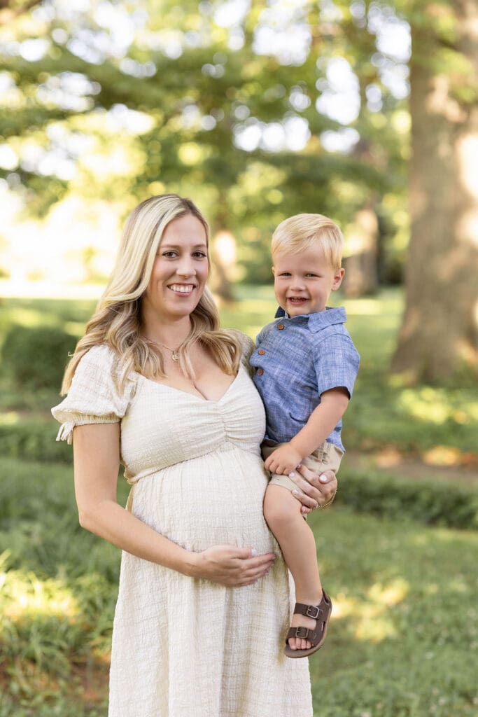 family maternity photography Louisville KY pregnant mother in white dress smiling while holding toddler son outdoors under large tree