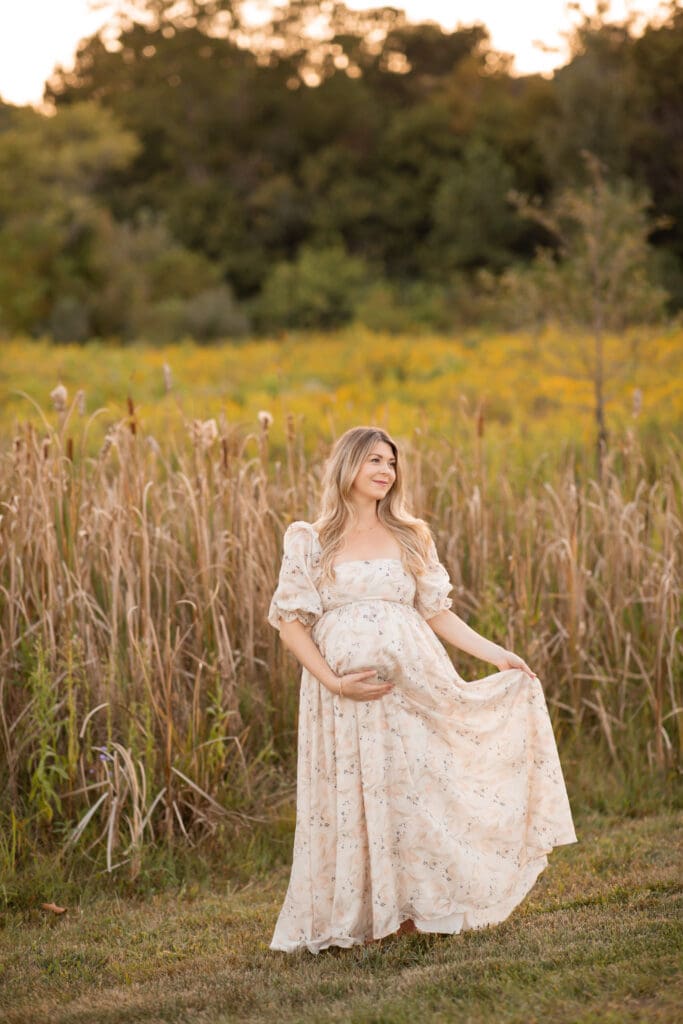 outdoor maternity photographer Louisville KY pregnant woman in cream floral dress twirling skirt in tall grass field at golden hour