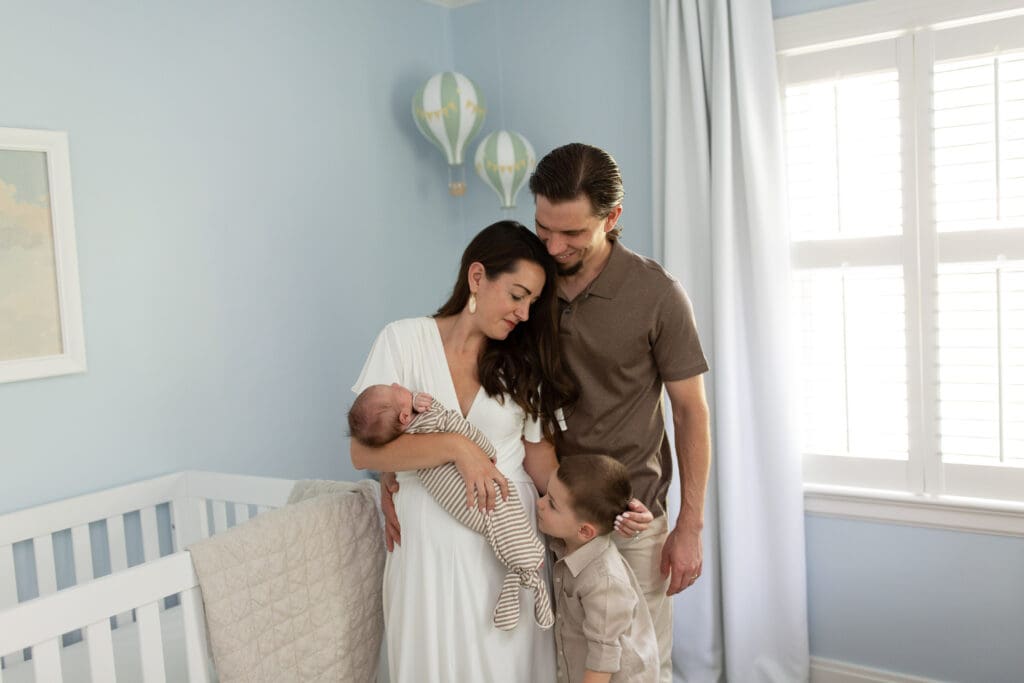 lifestyle newborn photography Louisville KY mother and father holding newborn baby in blue nursery with toddler son beside white crib