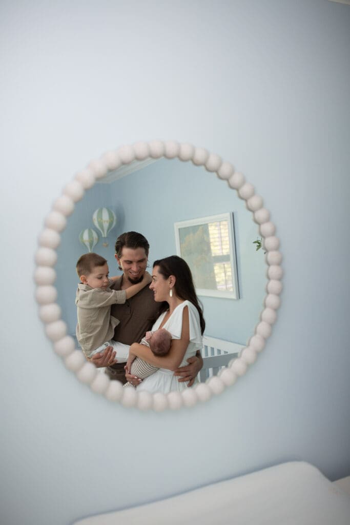 
in home newborn session Louisville KY family reflected in round nursery mirror with mother father toddler and newborn baby in blue room