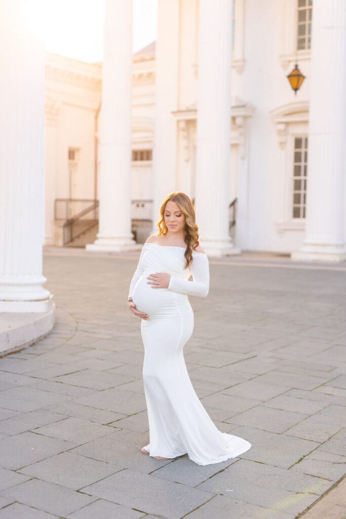 Expectant mother in flowing white gown cradling her baby bump at golden hour among the columns of the Louisville Water Tower in Kentucky