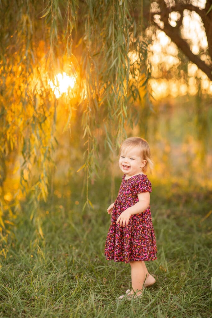 family maternity photography Louisville KY toddler girl in floral dress laughing under weeping willow tree at golden hour sunset