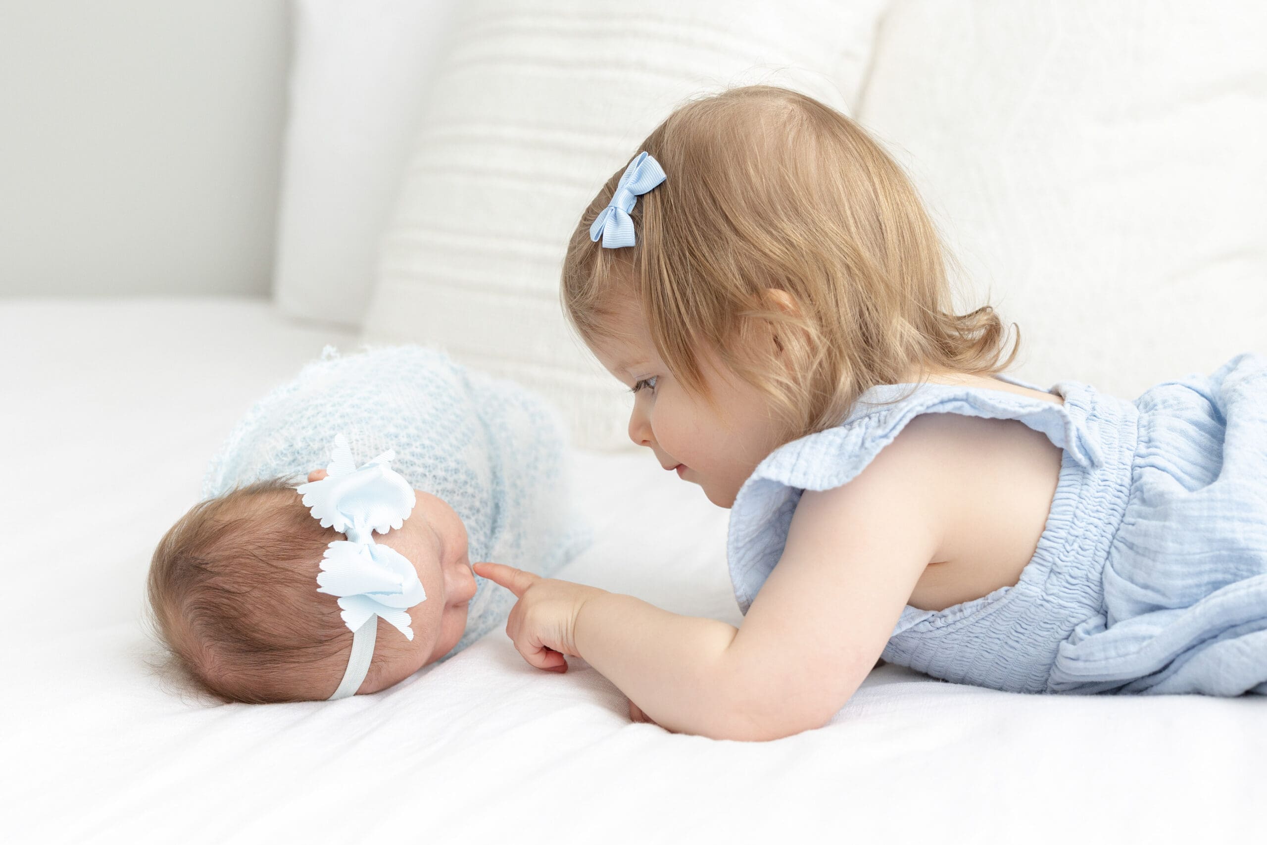 lifestyle newborn photography Louisville KY toddler girl in blue romper touching newborn baby sister wrapped in blue on white bed
