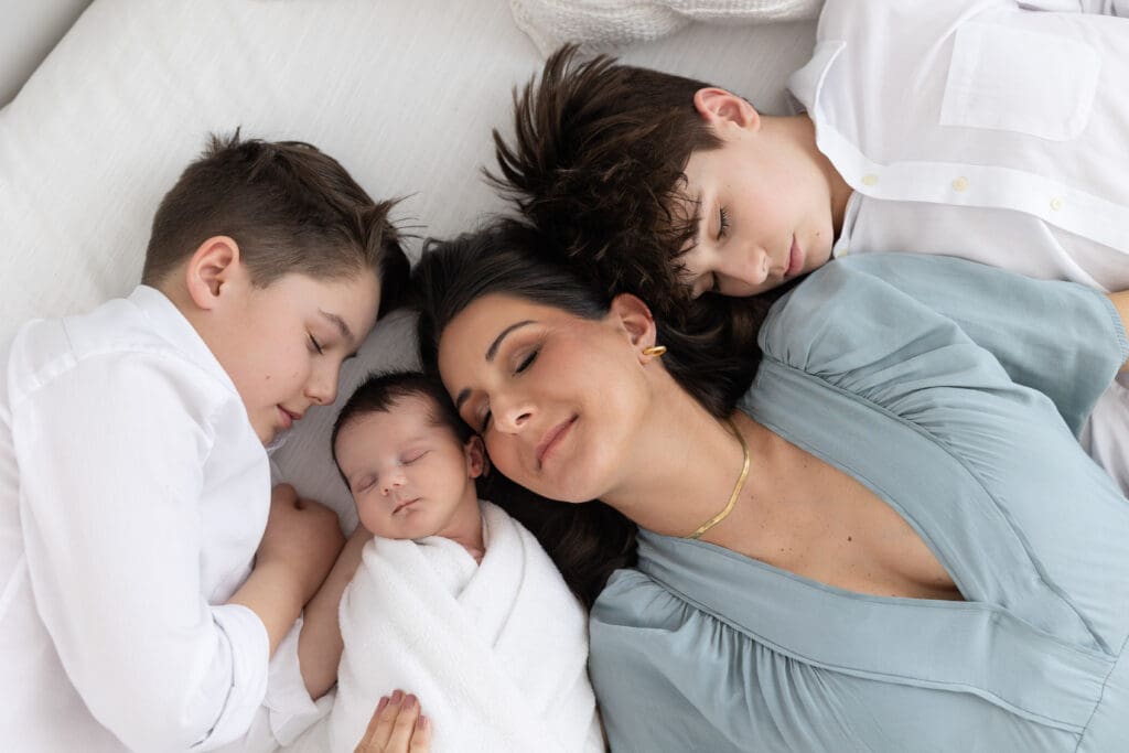lifestyle newborn photography Louisville KY mother lying on bed with newborn baby and two older sons in white and sage green