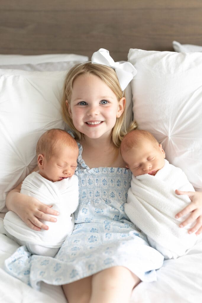 
in home newborn session Louisville KY big sister holding twin newborn babies on bed in blue and white outfits