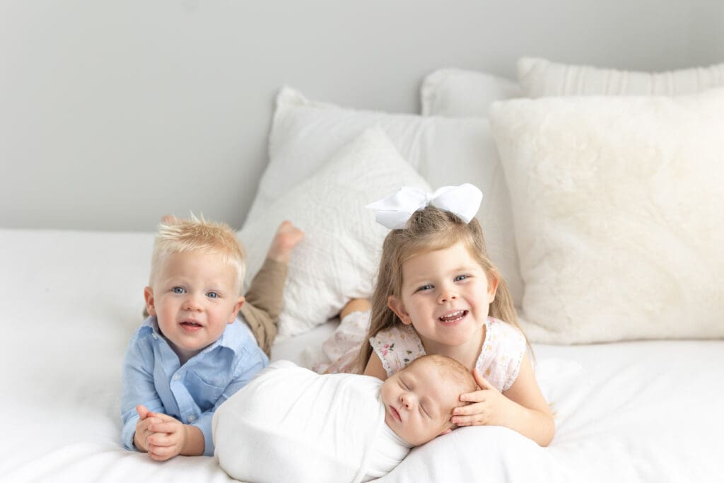 studio newborn photography Louisville KY toddler brother and older sister smiling with sleeping newborn baby wrapped in white on white bed