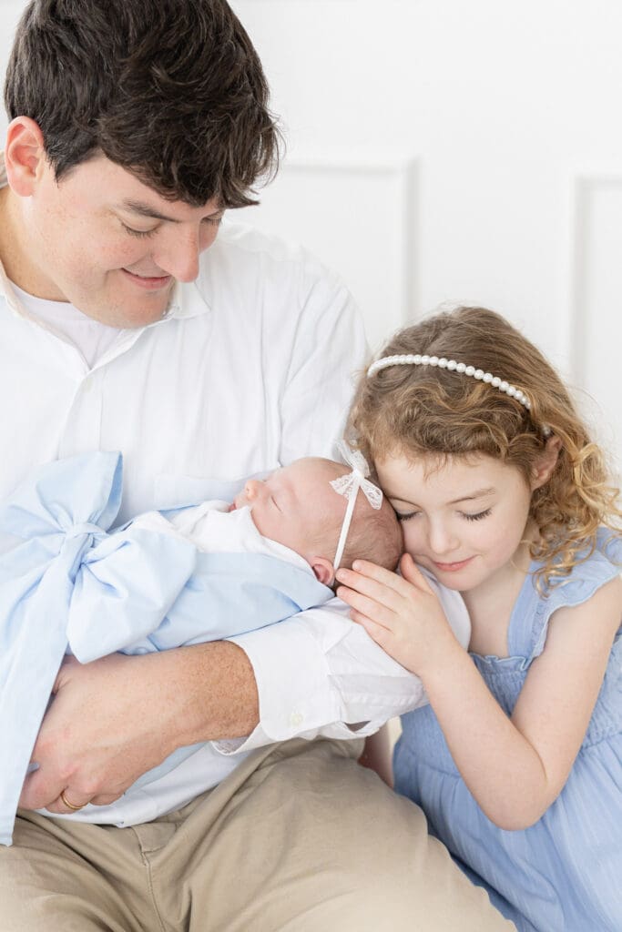 studio newborn photography Louisville KY father and older sister in blue dress cuddling sleeping newborn baby wrapped in blue on white bed
