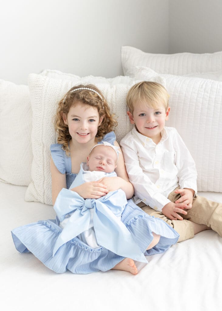 studio newborn session Louisville KY older sister and brother in blue outfits sitting on white bed holding newborn baby wrapped in blue bow blanket
