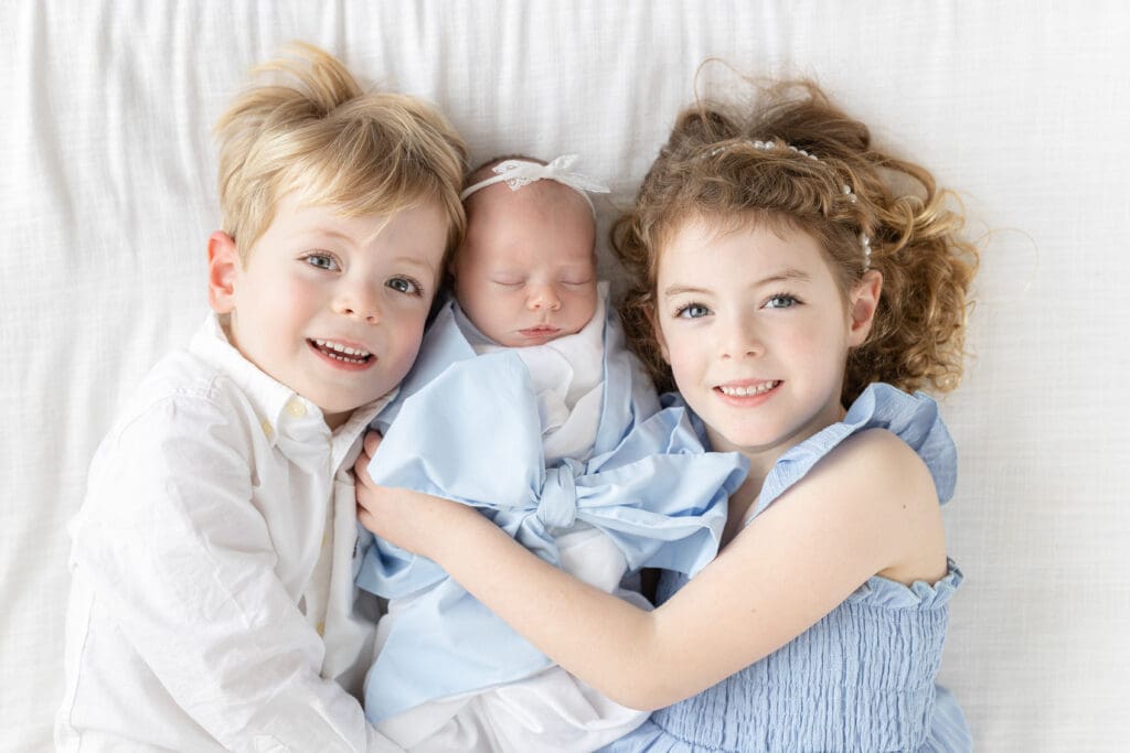 
studio newborn photography Louisville KY overhead shot of older brother and sister in blue smiling while cuddling newborn baby wrapped in blue bow on white bed