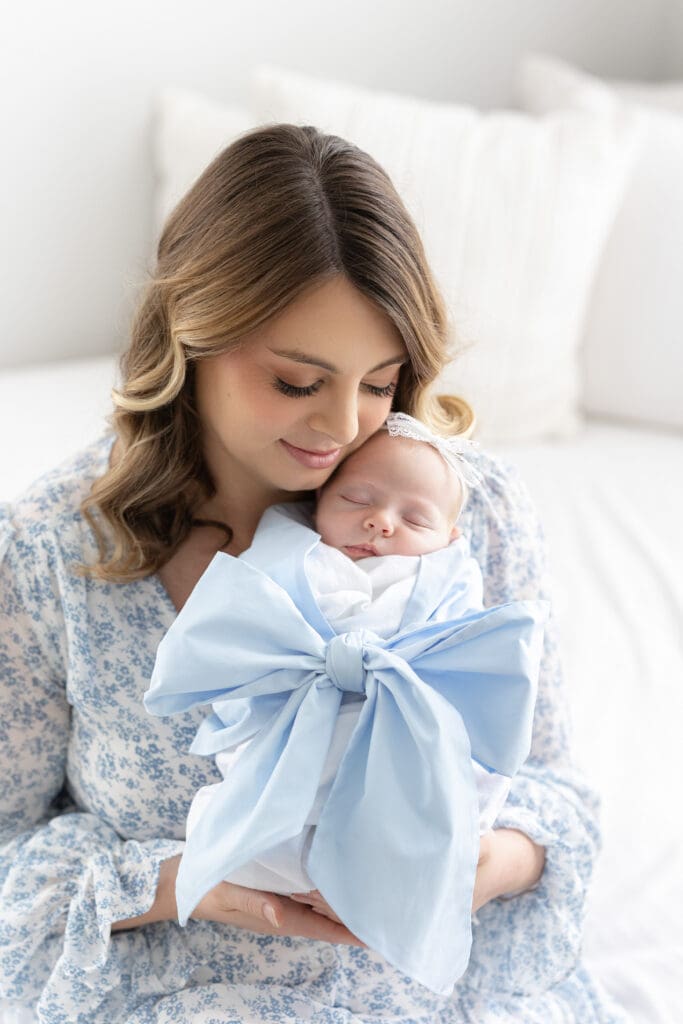 newborn photographer Louisville KY mother in floral blue dress holding sleeping newborn baby wrapped in blue bow against white background